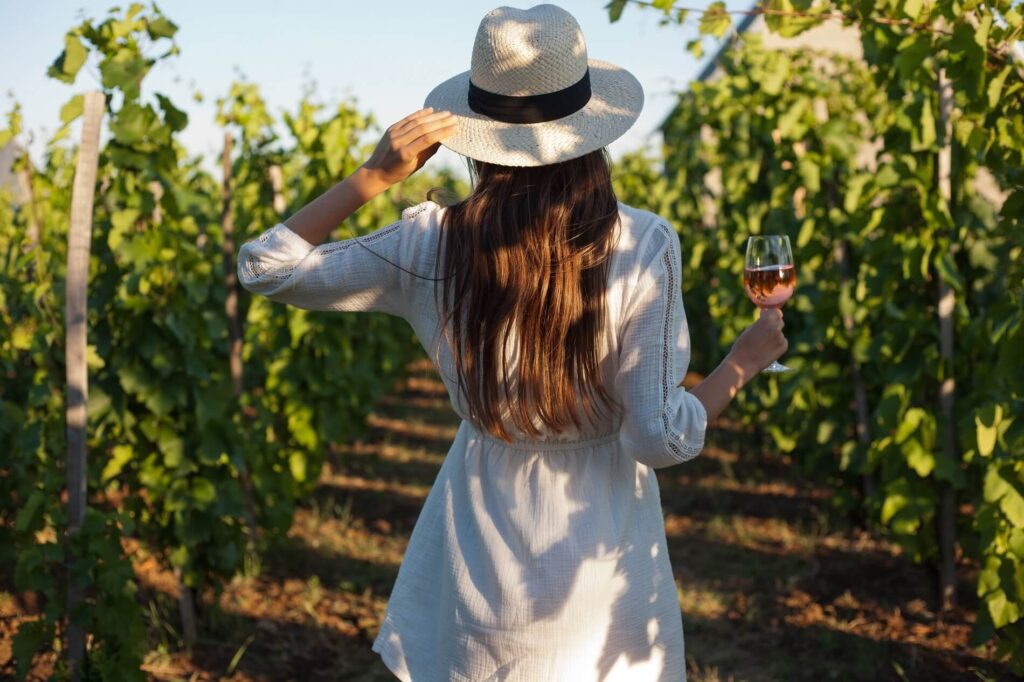 Une femme vue de dos, portant un chapeau et tenant un verre de vin, se promène dans un vignoble.