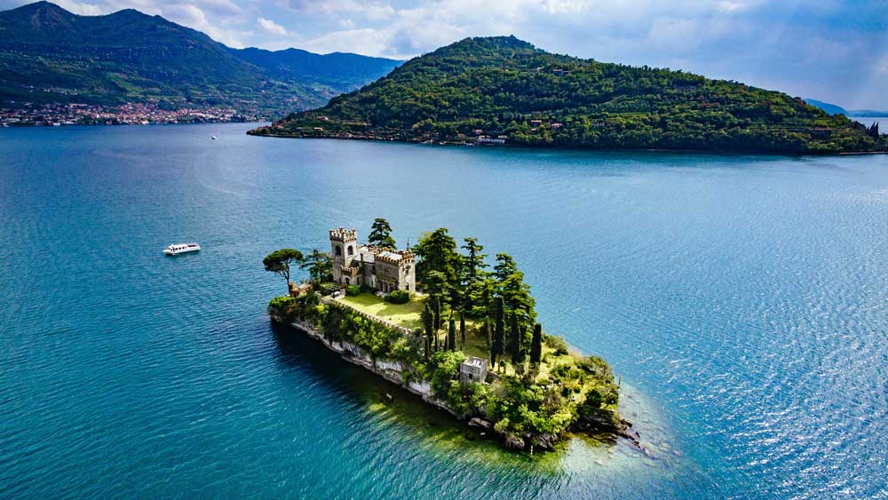 Isola con vegetazione rigogliosa e una piccola costruzione in un lago tranquillo, circondata da montagne verdi.